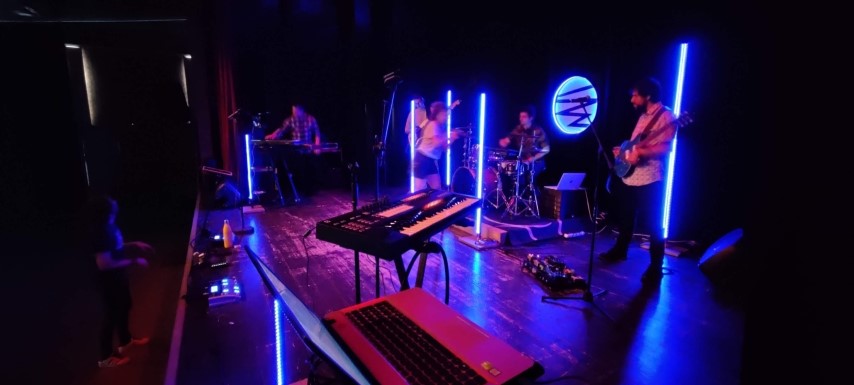 La banda tocant a l’escenari, amb llum blava i tubs de neó.
